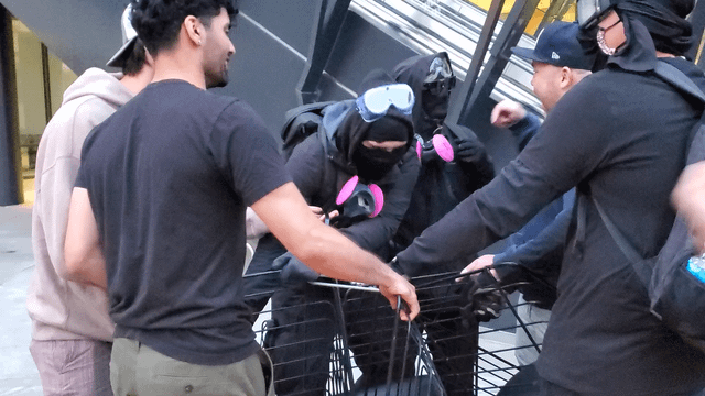 Blac block members and protesters wrestle over outdoor chairs from Wells Fargo Center, ultimately dragging them to the street