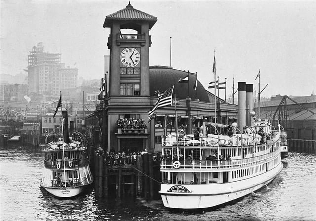 View of Colman Dock, Pier 52, now the Washington State Ferries terminal, with 2 mosquito fleet ferries tied up and the Smith Tower undergoing construction in the background. c. 1911.